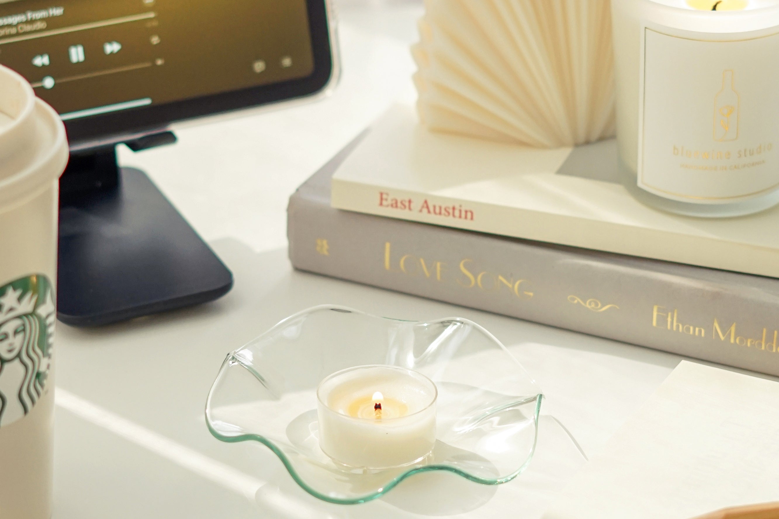 a lit tealight candle on a clear wavy ruffle dish, starbucks tumbler, pencil , and a lit frosted glass container soy candle on books