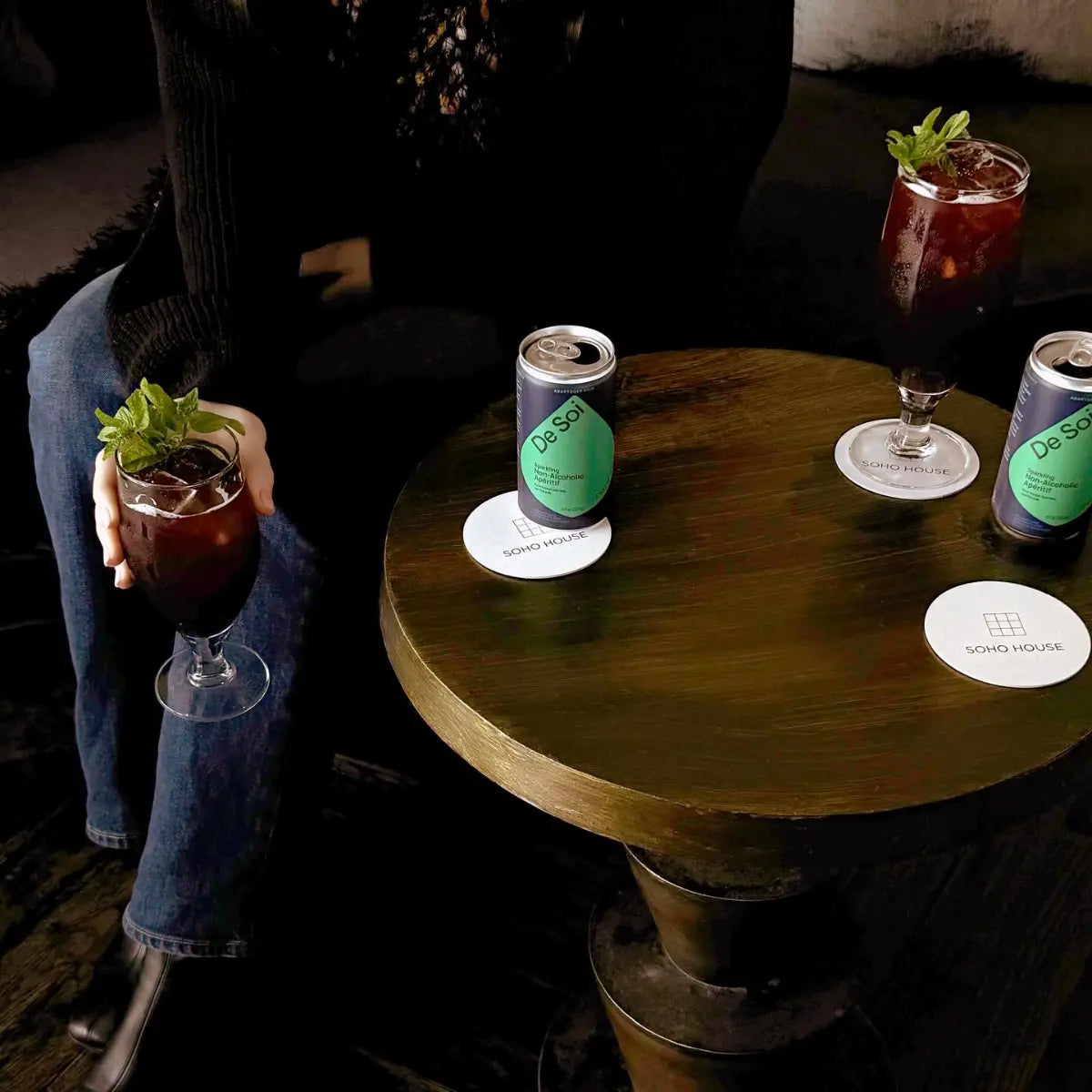 Adaptogen mocktails served in stemmed glasses with mint garnish alongside De Soi cans on a dimly lit lounge table.