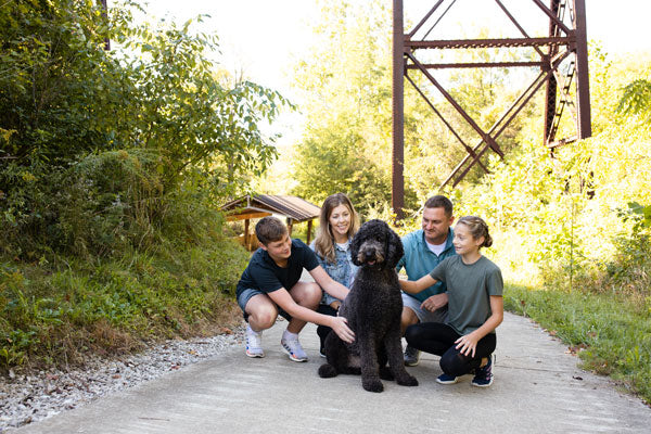 family of four kneeling and petting a black dog on an outdoor trail
