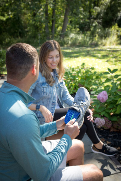 man sitting on a bench next to a woman while placing a blue insole into a gray shoe