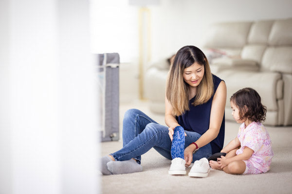 mother placing blue insole into white shoe sitting on floor with daughter