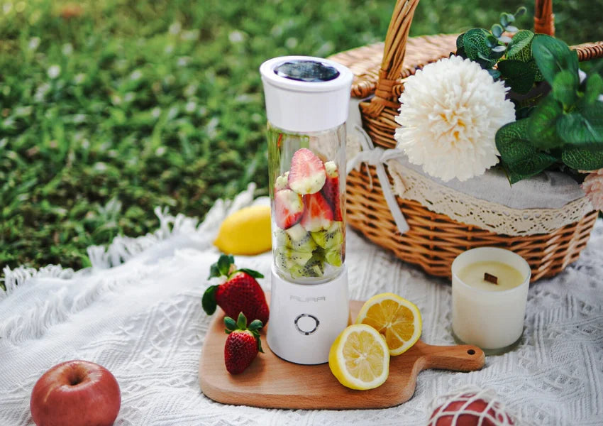 aura portable blender with fruits like sliced lemon, strawberries, apples and kiwi at a picnic