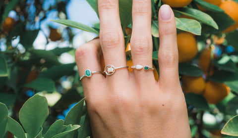 Hand mit drei Ringen aus Silber mit Smaragden und einem Opal vor einem Orangen Baum