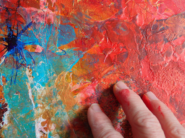 Kimberli Werner's hand touching an abstract painting made with red, orange, blue, and turquoise paints.