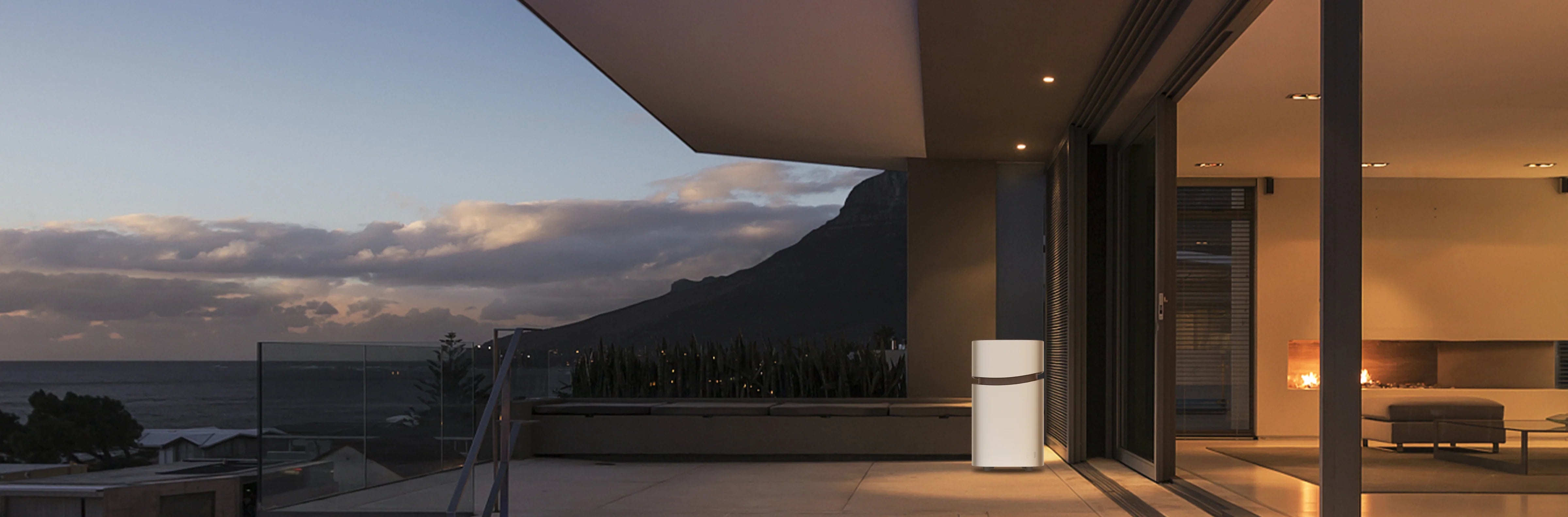 A modern patio at sunset with mountain and sea views, featuring large glass doors, outdoor seating, and a Mango Power 4,350W 6,900Wh Union Portable Battery and Power Station by Mango Power near the entrance for seamless backup power.