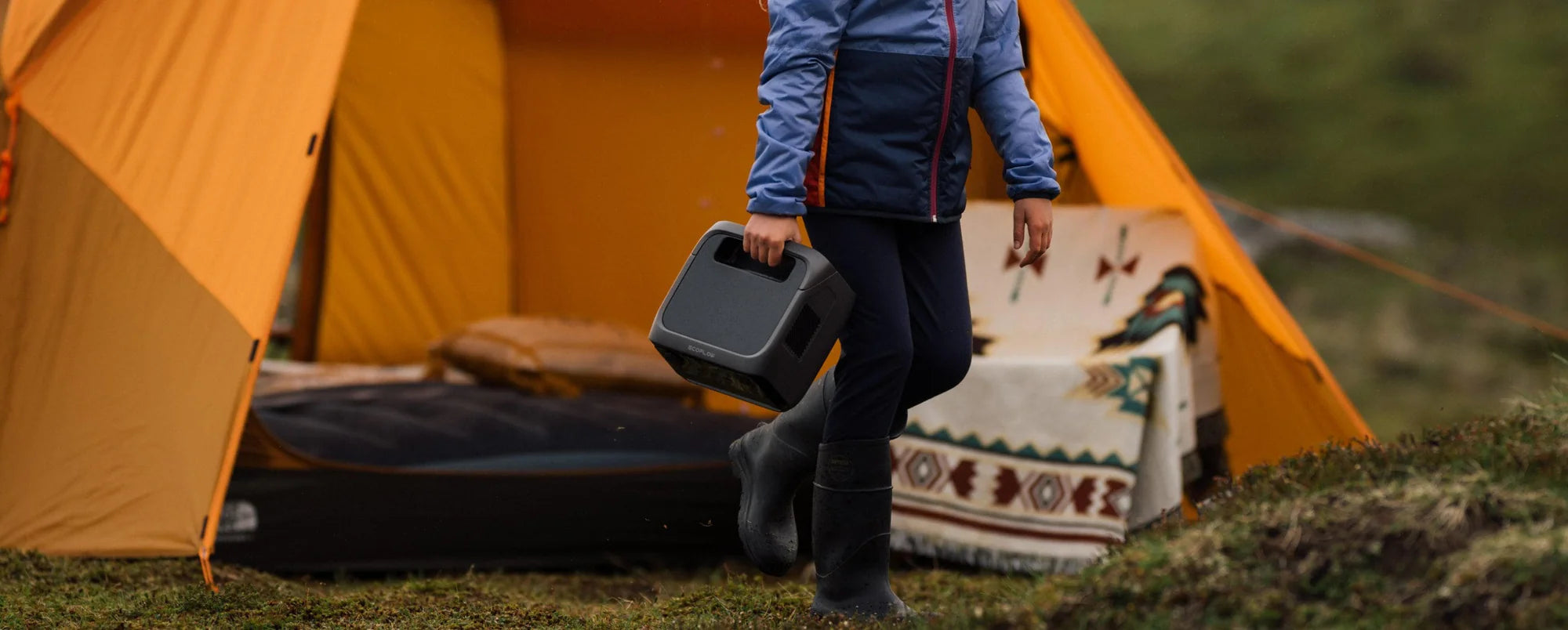 A person in a blue jacket and black boots carries an EcoFlow RIVER 3 Plus 600W 286Wh Portable Solar Power Station outside a yellow camping tent on grass, with a patterned blanket and air mattress inside the tent.