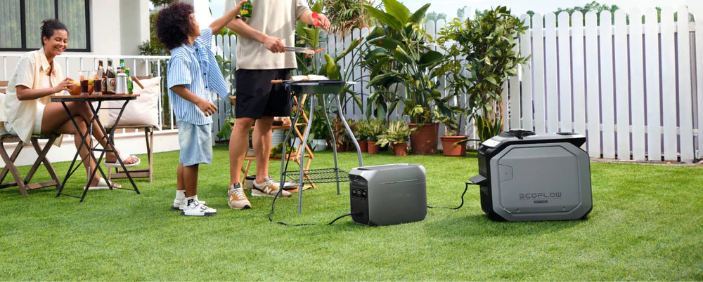 A family enjoys a backyard barbecue as a woman sits at a table, while a man and child grill food powered by the EcoFlow DELTA 3 Plus 1,800W 1,024Wh Portable Solar Power Station. Potted plants and a white fence are in the background.