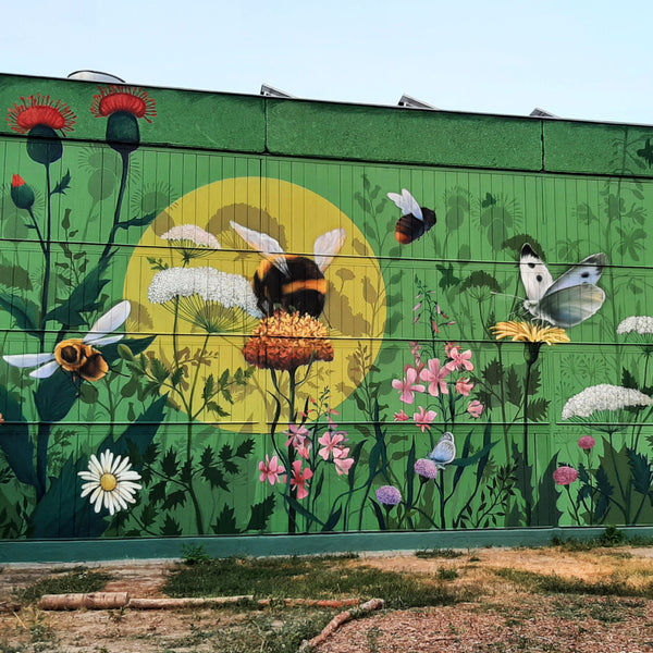 Photo showing a large mural on the side of an industrial building, depicting wildflowers swarmed by bumblebees and other pollinators like butterflies and other insects.