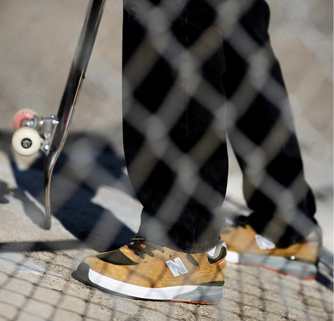 Andrew Reynolds wearing New Balance Numeric 933 skate shoes with skateboard, viewed through chain-link fence at a skate spot.