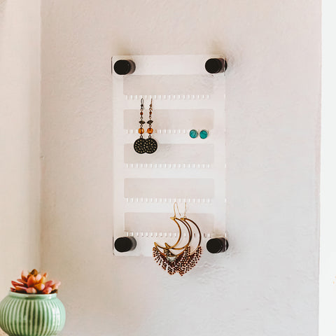 6 X 10 Clear acrylic earring holder hanging on the wall with several pairs of earrings hanging on it.
