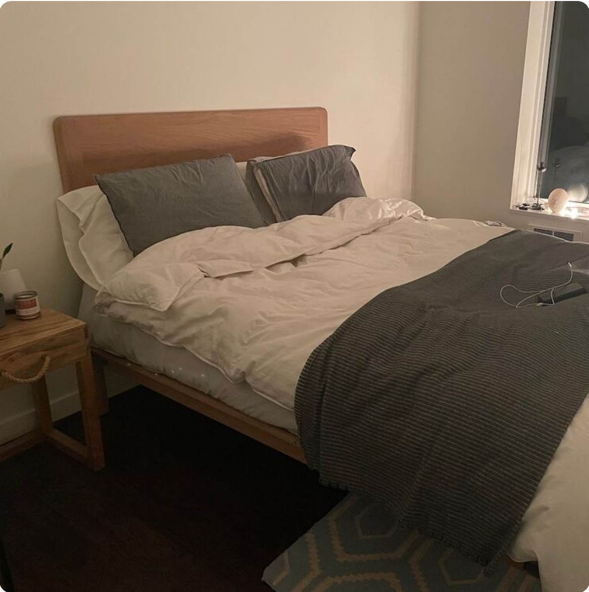 A neatly made bed with white sheets and gray pillows, beside a wooden nightstand.