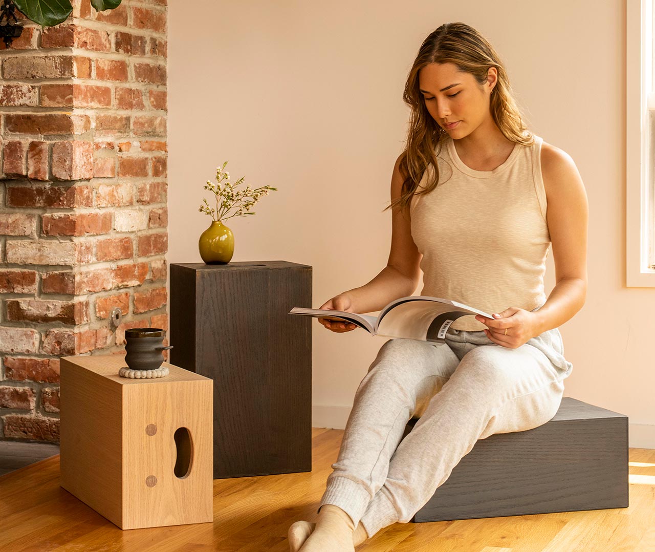 Woman reading a magazine while sitting near a brick wall and minimalistic furniture.