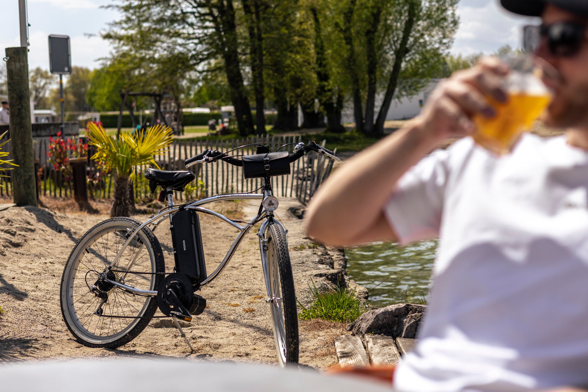 Homme dégustant une bière sur la plage, avec un vélo de plage de couleur sable amélioré par un kit de conversion de vélo électrique RocketBike X1 Photon, en arrière-plan - solution de cyclisme durable et économique.