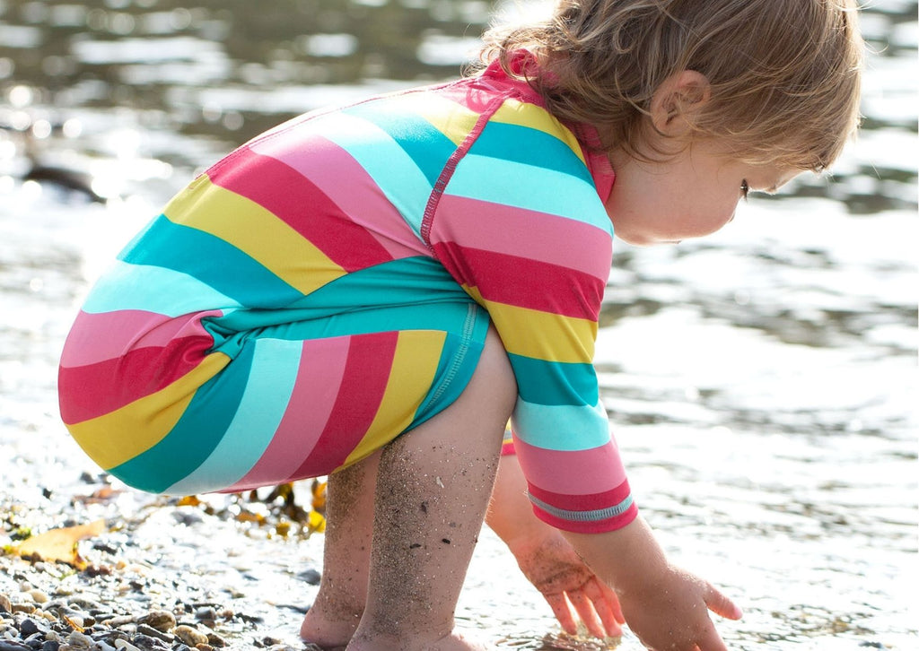 A little girl crouching near water wearing a Frugi toddler surfsuit