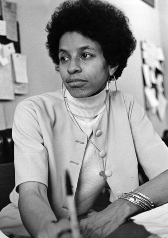 A black and white photo of poet Dr. Johari M Amini-Hudson, a black woman wearing a light grey cardigan, sitting  in a chair. She has a medium msized afro, her hair is black, against a light coloured background
