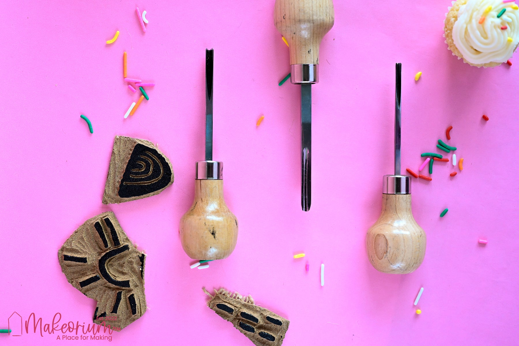 Three linocut cutting tools with wooden handles and linocut stamps on a bright pink background surrounded by rainbow jimmies sprinkles