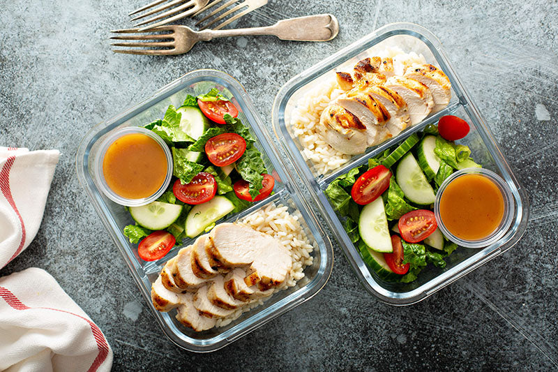 Aerial view of two meal containers filled with a healthy combination: Fresh salad with tomatoes, cucumbers, and dressing alongside grilled chicken and rice.