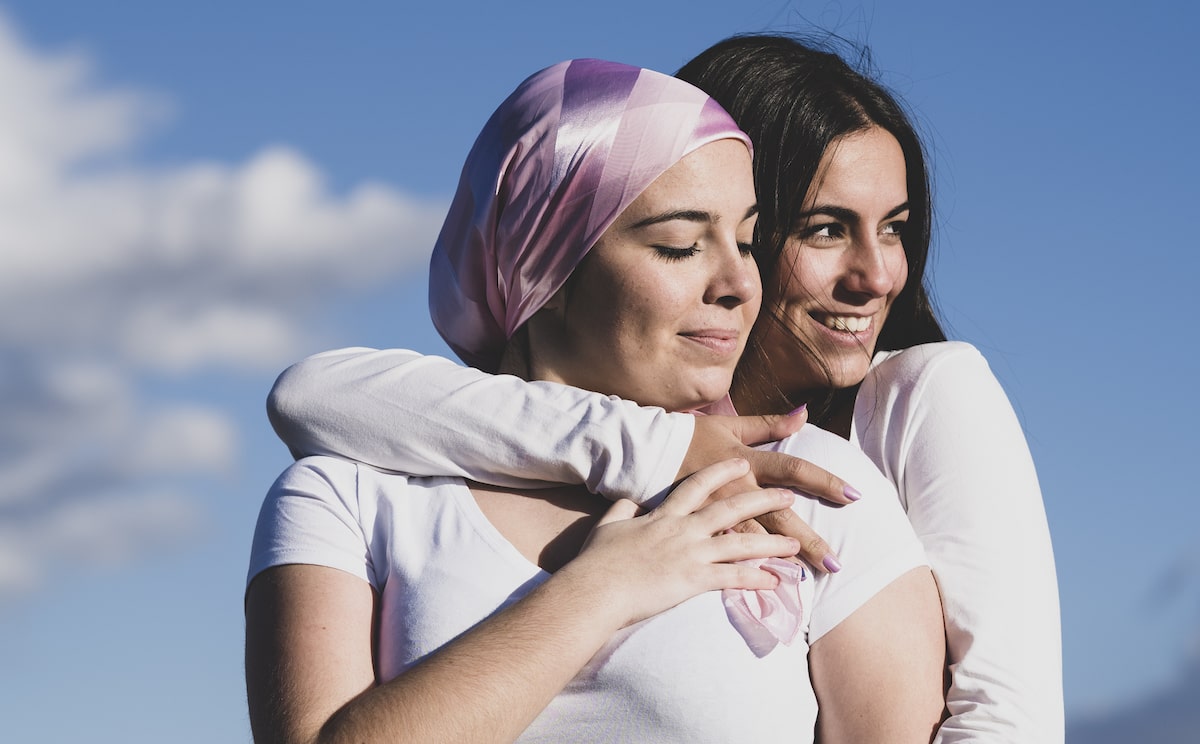 Two women hugging holding a pink ribbon and one wearing a pink bandana
