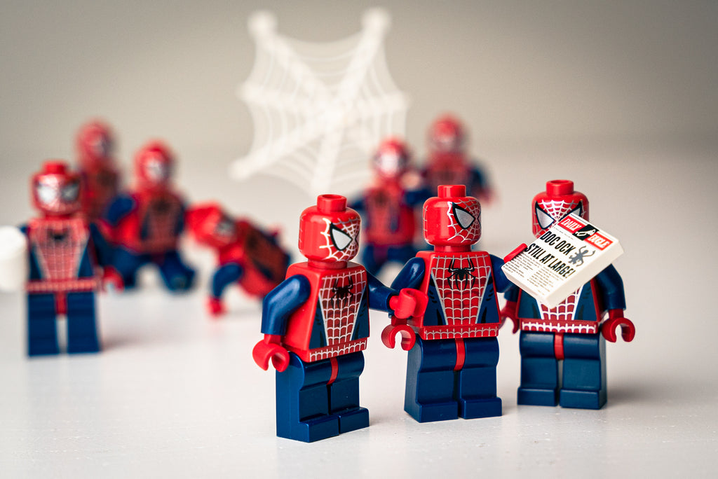 Image of Spider-Man Lego minifigures: A creative arrangement of multiple Spider-Man Lego minifigures in various poses, with a focused foreground and a blurry background. One figure is highlighted holding a newspaper with the headline "DOCK OCK STILL AT LARGE."