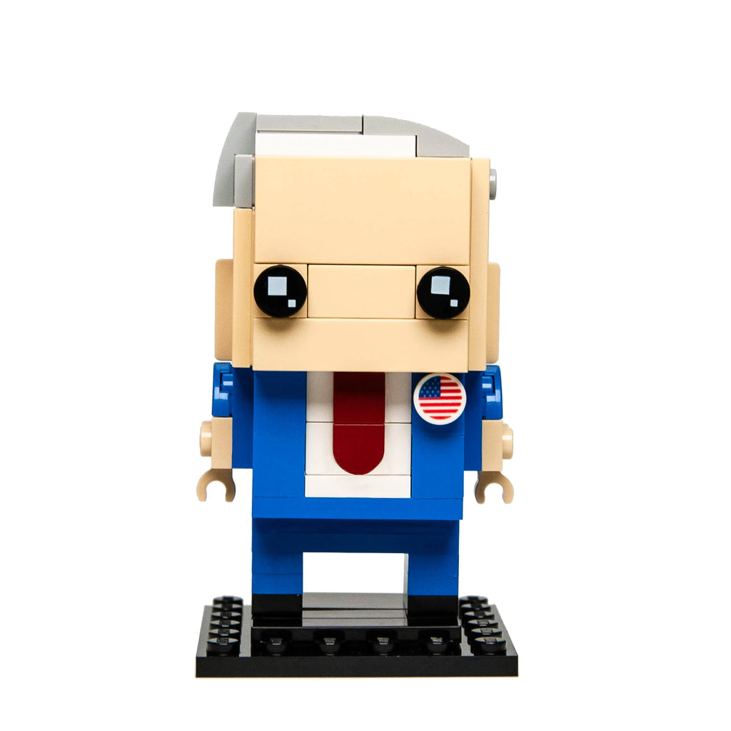 Custom Lego BrickHeadz figure of President Biden with gray hair, blue suit, red tie, and American flag pin, presented on a white background.