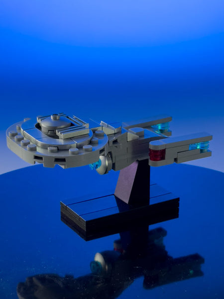 A sophisticated Lego model of a spaceship stands mounted on a sleek black stand, set against a gradient blue background that fades from a deep sea blue at the top to a lighter sky blue. The spaceship, designed with intricate gray bricks, features a circular array of blocks resembling a ring around the main body, highlighted with translucent blue and red pieces suggesting lights or sensors.