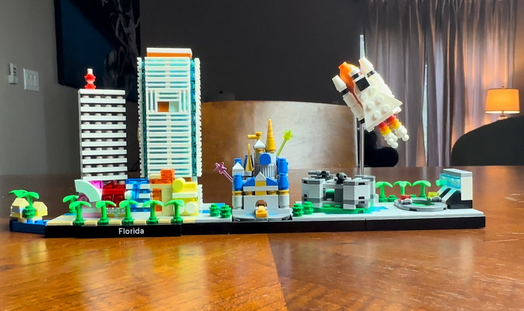 A vibrant Lego cityscape sprawls across a flat surface, encapsulating the essence of Florida with miniature representations of iconic structures and a rocket mid-launch. The colorful display includes palm trees, a high-rise building with a red beacon on top, a stylized castle with playful star and flower details, and a white rocket with orange and red thrusters, all set on a base that proudly declares 'Florida' in bold lettering, inviting onlookers to a playful journey through a Lego lover's rendition of the Sunshine State