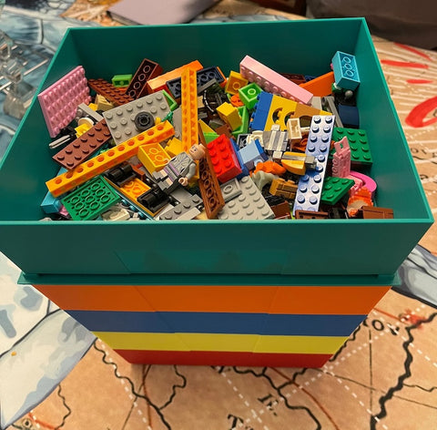 A colorful, teal-green LEGO storage box overflowing with an assortment of LEGO bricks and figurines in various colors and shapes, placed on a patterned surface.