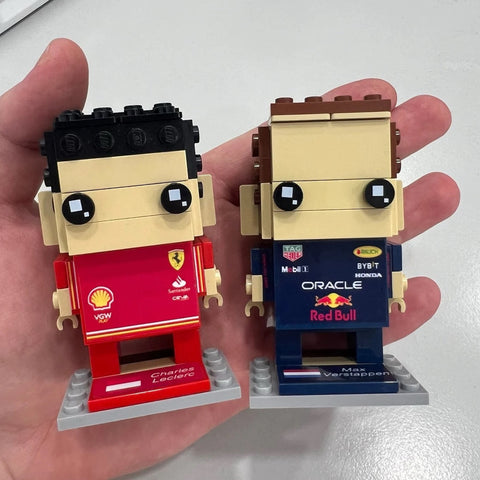 Two LEGO figures held in a hand, representing Formula 1 drivers Charles Leclerc and Max Verstappen, with team and sponsor logos on their outfits.