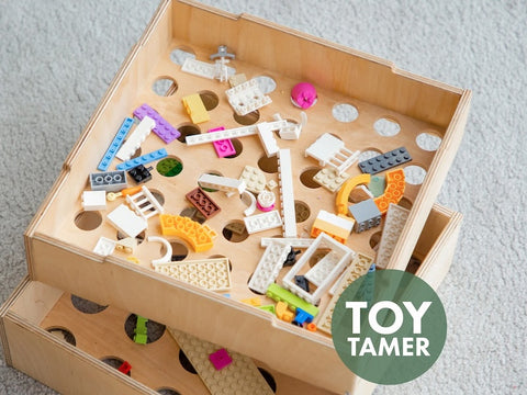 A wooden storage box filled with various colored and shaped LEGO pieces, including bricks, blocks, and connectors, on a carpeted floor. A label reading "TOY TAMER" is visible.