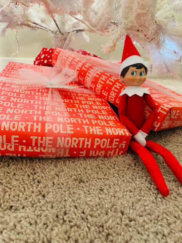 an Elf on the Shelf Scout Elf sitting under a flocked Christmas Tree with two presents from the North Pole