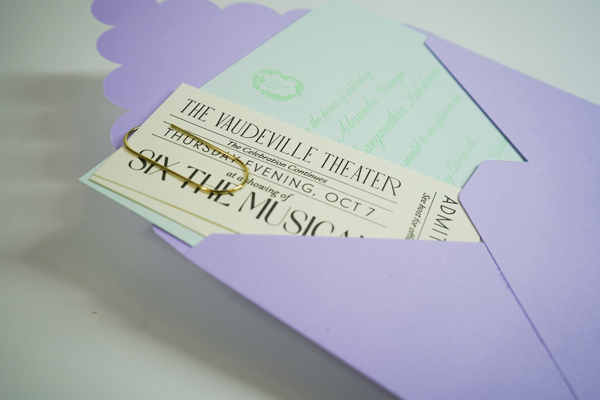 Scalloped envelope holding letterpress tickets and invitation, held together with gold wide paperclip