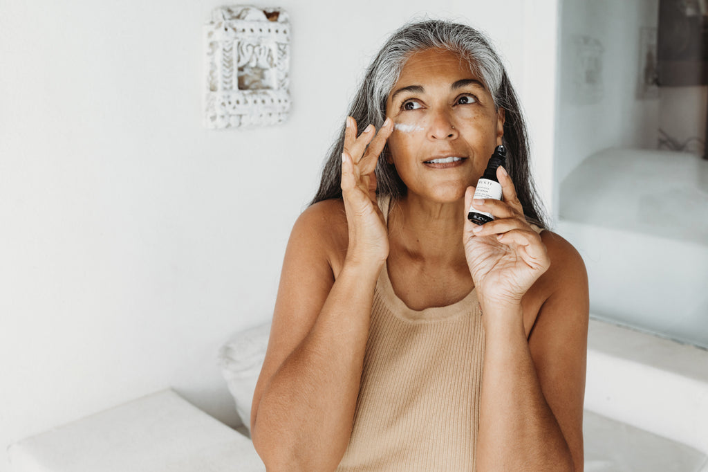 a beautiful woman with long salt and pepper hair wears a beige top and holds a small bottle up to her face, with the other hand she is applying eye serum