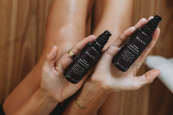 a pair of hands hold two bottles of Mukti's moisturisers with sunscreen, tinted and daily, over their knees and in the background is a wooden floor.