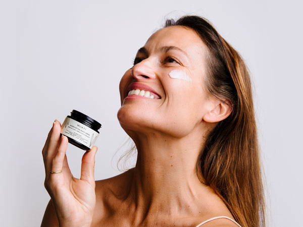 A woman with long brown hair is looking up and smiling, she has a swipe of product on her cheek. her hand is near her face holding an open jar of the product, Queen of the Night Crème.