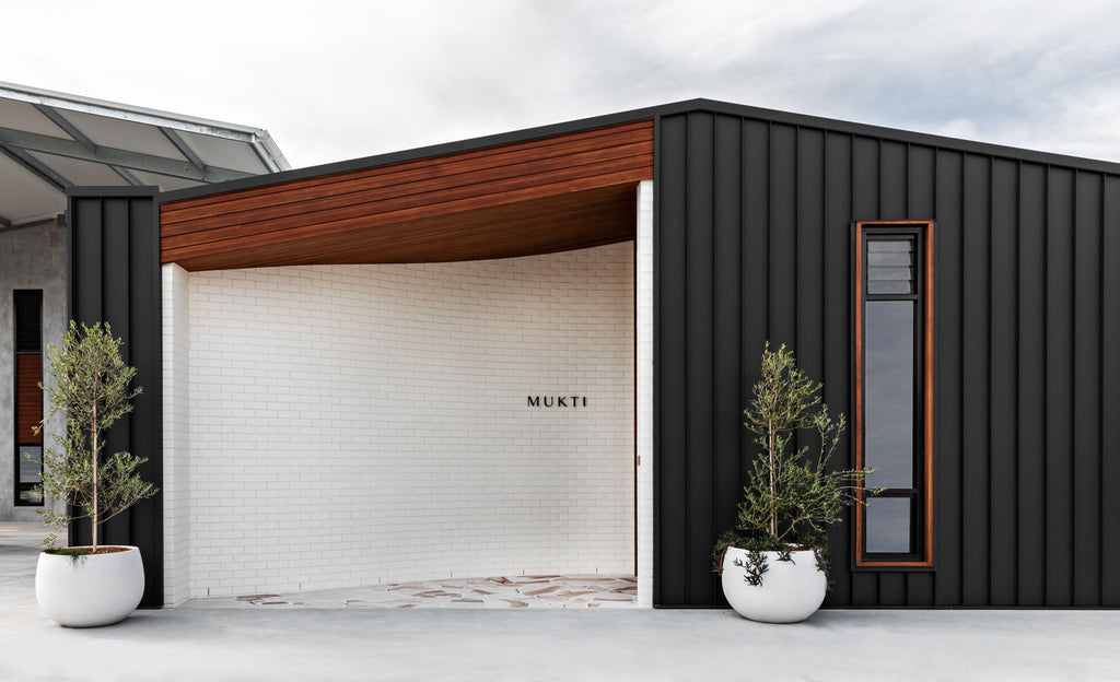 a beautiful architecturally designed building with dark wood, black steel and cream brickwork above a natural-toned stonework floor, two olive trees in white round pots frame the entrance