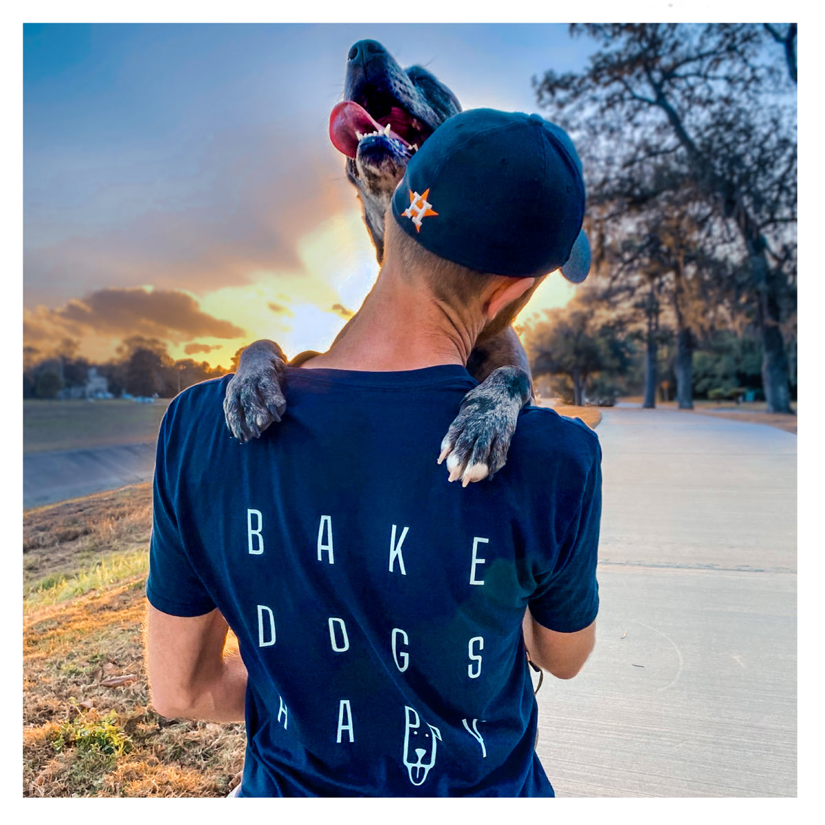 Photo of a large black and grey dog hugging one of the BakedBones founders who is wearing a Houston Astros baseball cap and a t-shirt that says 