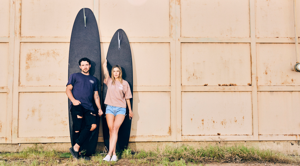 Man and Women leaning up against a wall with a surfboard wearing Omeek brand tee shirts