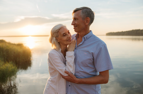 An older couple smiling and posing together as the sun sets over the body of water behind the couple.