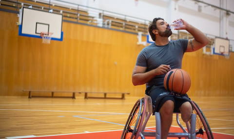 A man in a wheelchair drinks water after playing basketball on a court.