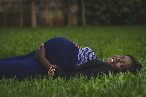 Pregnant woman preventing kidney stones in blue dress lies on green grass, eyes closed with hands on baby bump, surrounded by trees and blue sky with clouds