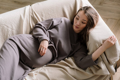 Pregnant woman preventing kidney stones in white dress lies on couch, with eyes looking at baby bump and hands on her stomach.