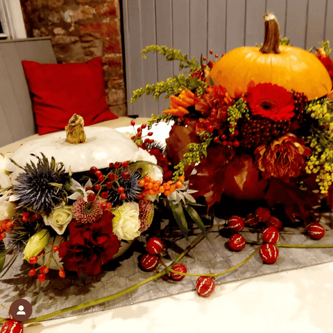 Photo showing a statement autumnal table flower arrangement. The arrangement is a white and an orange pumpkin. Both have flowers pouring out of their centre.