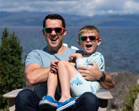 A man and a boy sitting by a lake wearing Sunnies shades polarized sunglasses with 100% UVA/UVB protection in the Sunnies Smash Up color