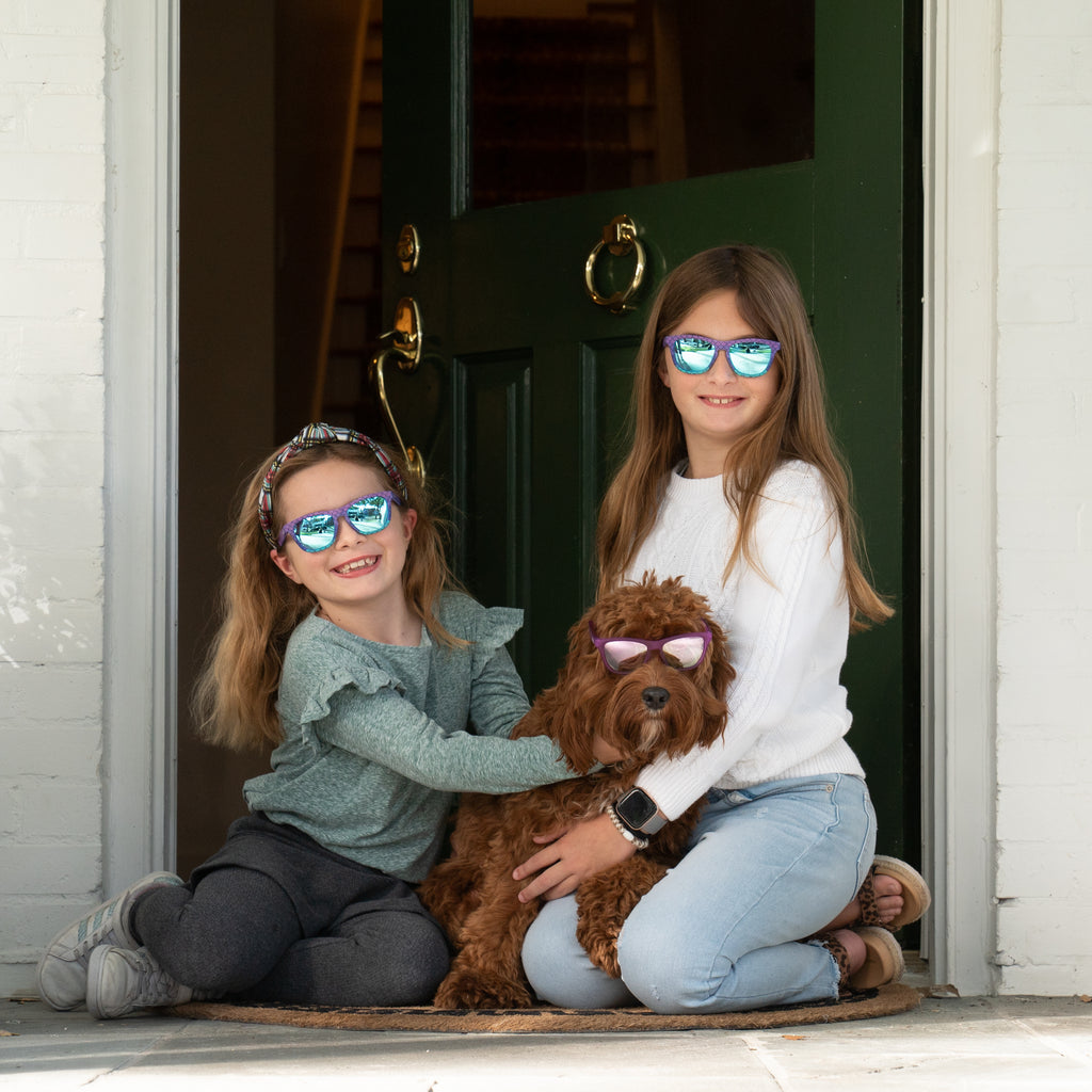 Two girls sitting on their front porch with their labradoodle wearing polarized kids sunglasses by Sunnies Shades.  The pup is also wearing Sunnies polarized kids sunglasses.