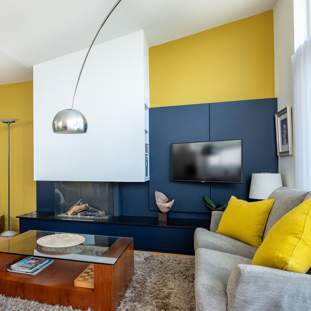 Living room with gold wall and dark navy tv unit, silver lamp, dark wooden coffee table, grey couch with yellow cushions