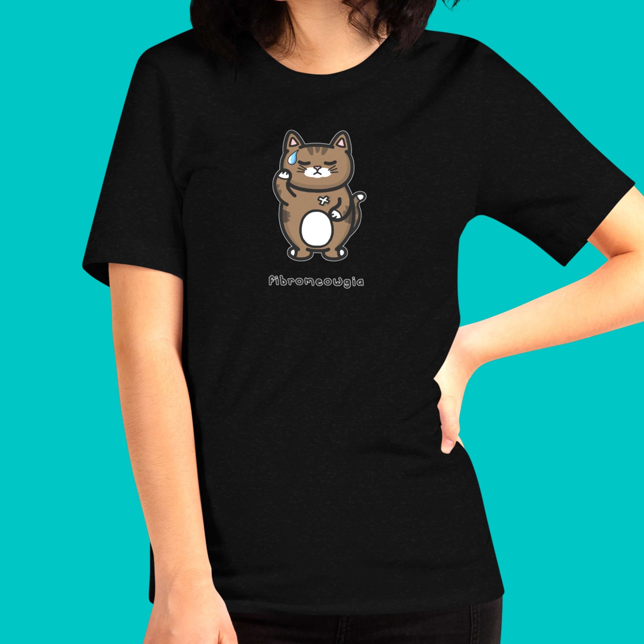 The Fibromeowgia Cat Tee - Fibromyalgia Syndrome modelled by a femme person with brown hair on a blue background. The black short sleeve tshirt features a sad brown tabby cat with a sweat droplet clutching its body with a white bandaid and bottom text reading 'fibromeowgia'. The hand drawn design is raising awareness for fibro fibromyalgia syndrome.