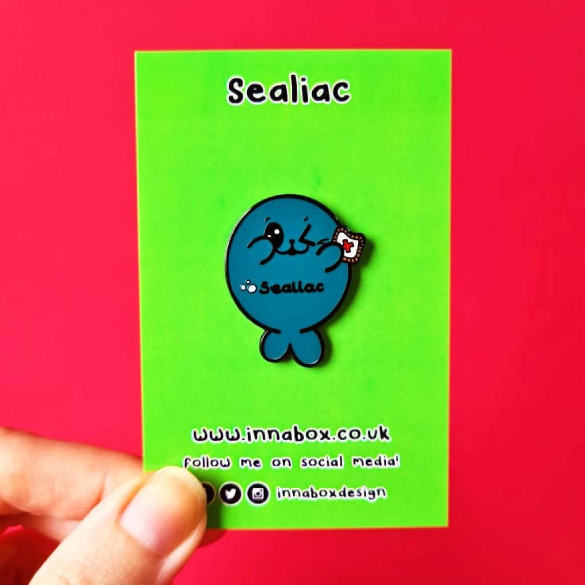The Sealiac Seal Enamel Pin - Coeliac Disease on green backing card held over a red background. A blue winking smiling seal holding up a piece of toast with a red cross, across its belly in black text reads 'sealiac'. The hand drawn design is raising awareness for coeliac disease.