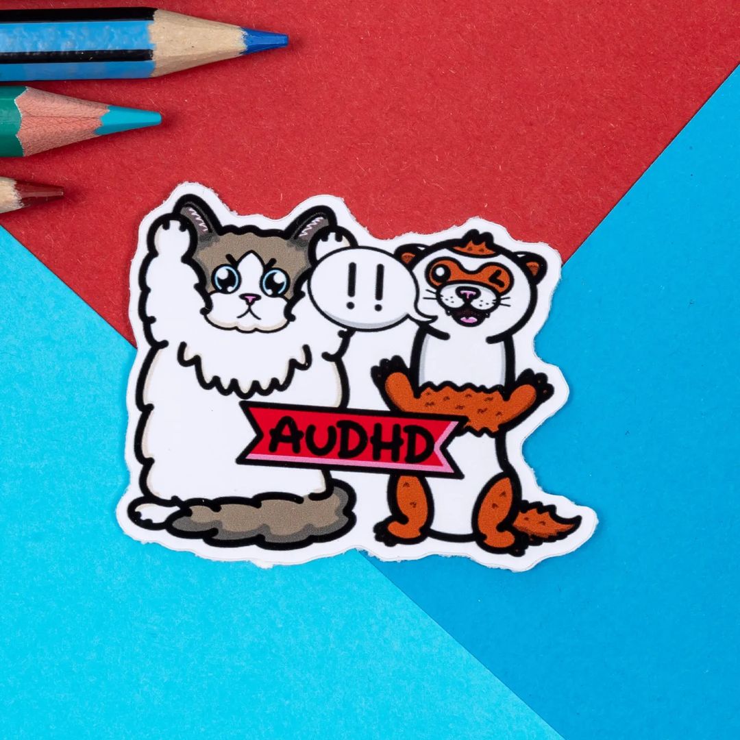 A ragdoll cat and ferret sticker on a blue and red background with colouring pencils. The cat on the left is clutching its ears with an angry expression, the ferret on the right is smiling and winking with a speech bubble '!!'. Across both characters is a red banner with black text reading 'AuDHD'. The hand drawn design is raising awareness for autism, adhd and audhd neurodivergent people.