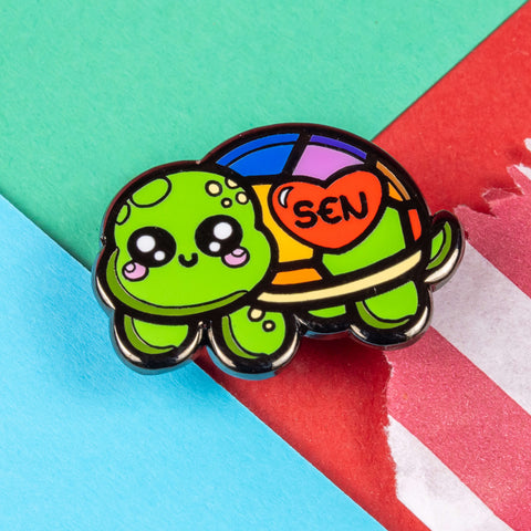 The Speshell Educational Needs Enamel Pin - SEN - Special Educational Needs on a red, blue and green card background. A rainbow shell kawaii cute style tortoise with pink cheeks and sparkling eyes, on its shell is a red heart with 'SEN' in the middle. The pin design is raising awareness for SEN Special Educational Needs.