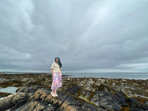 this is willow founder Lynda stands on some rocks in low tide at Skerries beach north of Dublin Ireland. A cloudy sky shows in an expanse behind and above. Lynda is wearing the Quinn Midi-Dress in pink with a cream 3/4 length sweater and pale pink shoes.